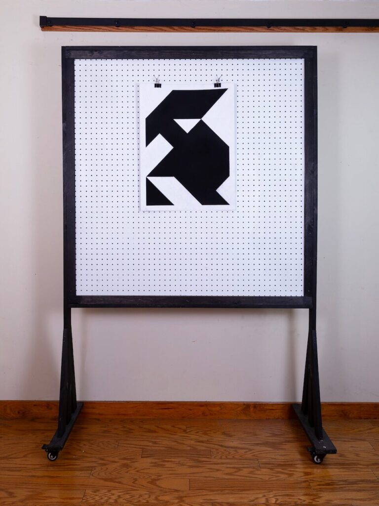 Caretaker (Glyph Drawing No. 5) displayed on studio pegboard, showing scale. Minimalist charcoal drawing with geometric forms and subtle figurative suggestion.