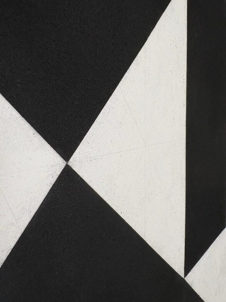 Detail of an abstract charcoal drawing, tightly controlled, showing a sandy, tactile texture, interrupted by a sharp white triangular shapes of the underlying paper.