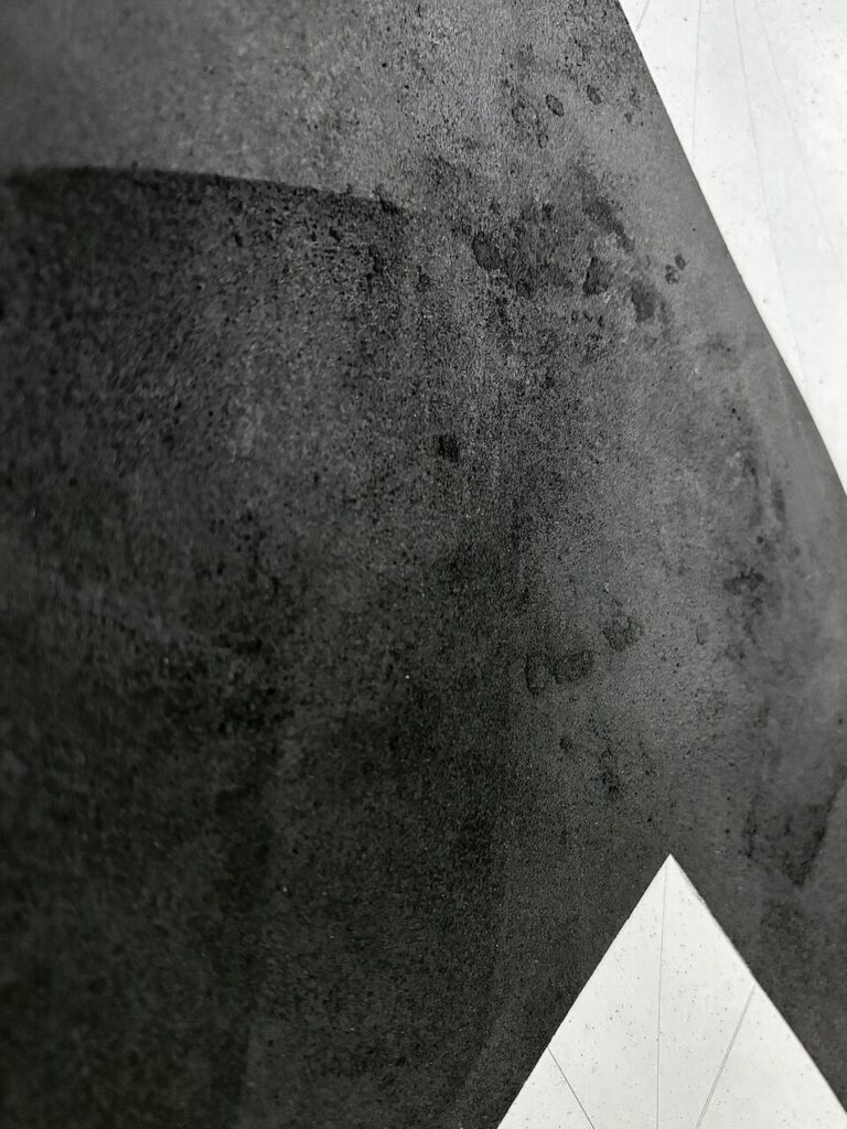 Detail of Mountain-Lodge (Glyph Drawing No. 9), showing a coarse, uneven surface, pockmarked and bearing the traces of brayers and scrapers. Charcoal has been worked on and off the surface reavealing oil stick beneath. The black is interrupted by a sharp white shapes of the underlying paper with faint graphite grid lines.