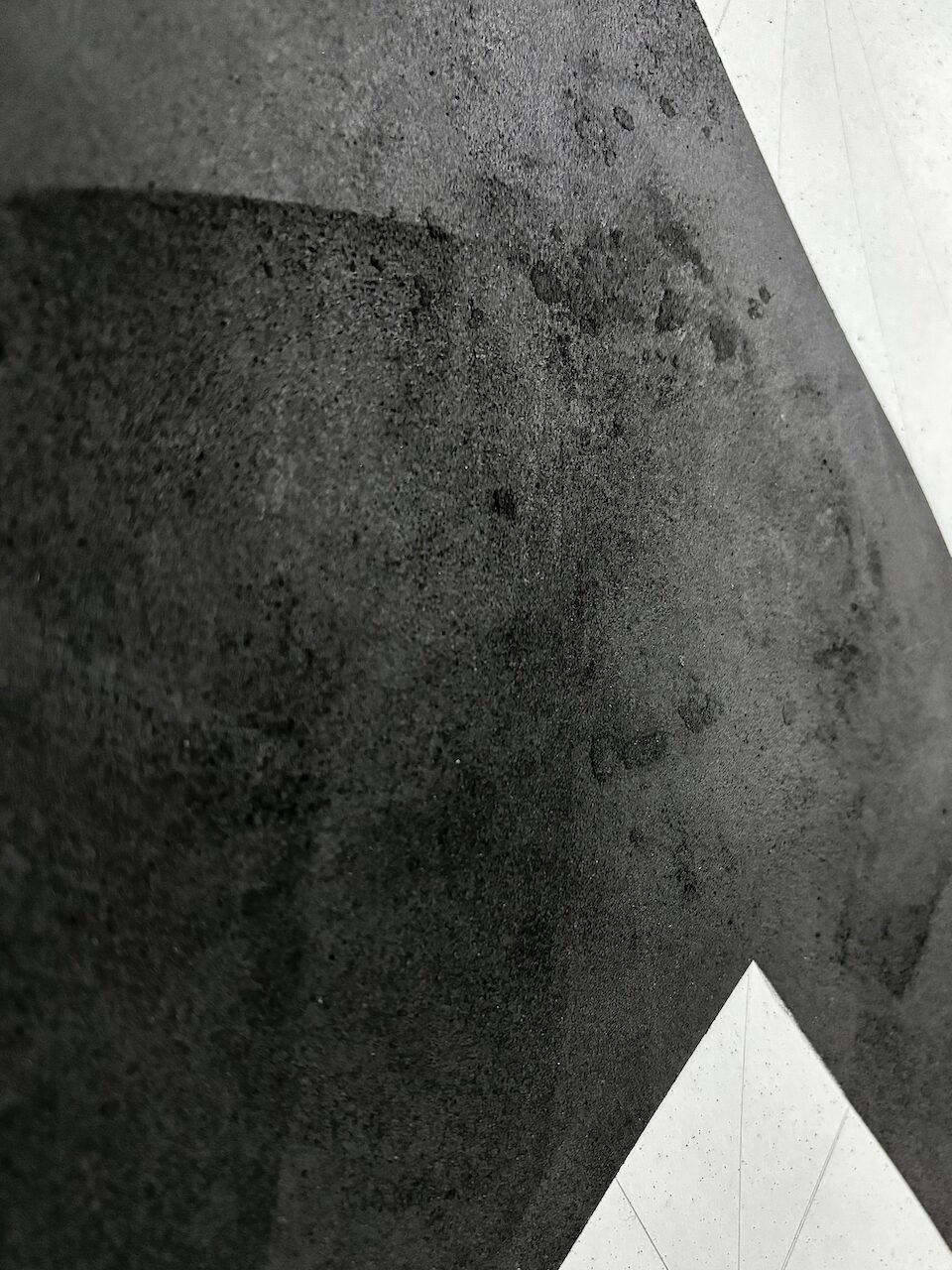 Detail of Mountain-Lodge (Drawing No. 12), showing a coarse, uneven surface, pockmarked and bearing the traces of brayers and scrapers. Charcoal has been worked on and off the surface reavealing oil stick beneath. The black is interrupted by a sharp white shapes of the underlying paper with faint graphite grid lines.