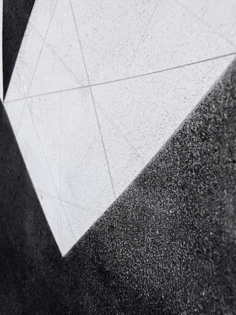 Detail of Neighborhood, from the Air (Glyph Drawing No. 7), showing an even, sandy, tactile black charcoal texture, black oil stick laid on by a brayer, interrupted by a sharp white shapes of the underlying paper with faint graphite grid lines.