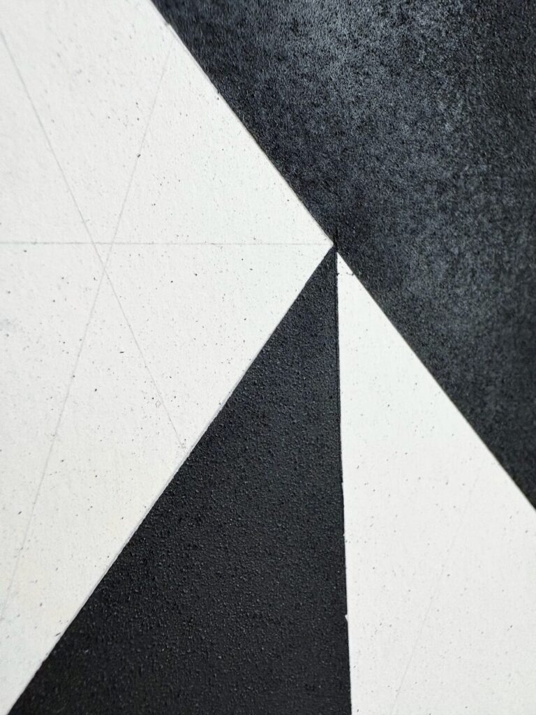 Detail of an abstract charcoal drawing, showing a sandy, tactile texture, interrupted by a sharp white triangular shapes of the underlying paper with faint graphite grid lines.