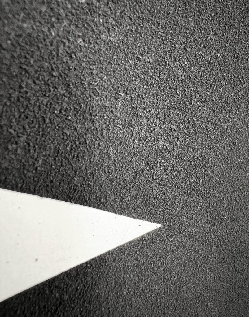 Detail of an abstract charcoal drawing, showing rich, even texture like fine sand, interrupted by a sharp white triangular cut into the surface.