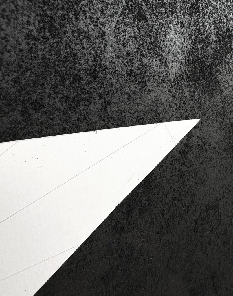 Detail of an abstract charcoal drawing, showing a distressed, pockmarked texture, interrupted by a sharp white triangular cut into the surface.