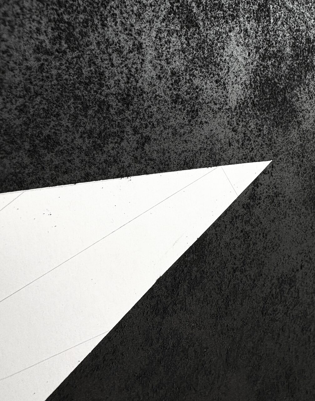 Detail of an abstract charcoal drawing, showing a distressed, pockmarked texture, interrupted by a sharp white triangular cut into the surface.