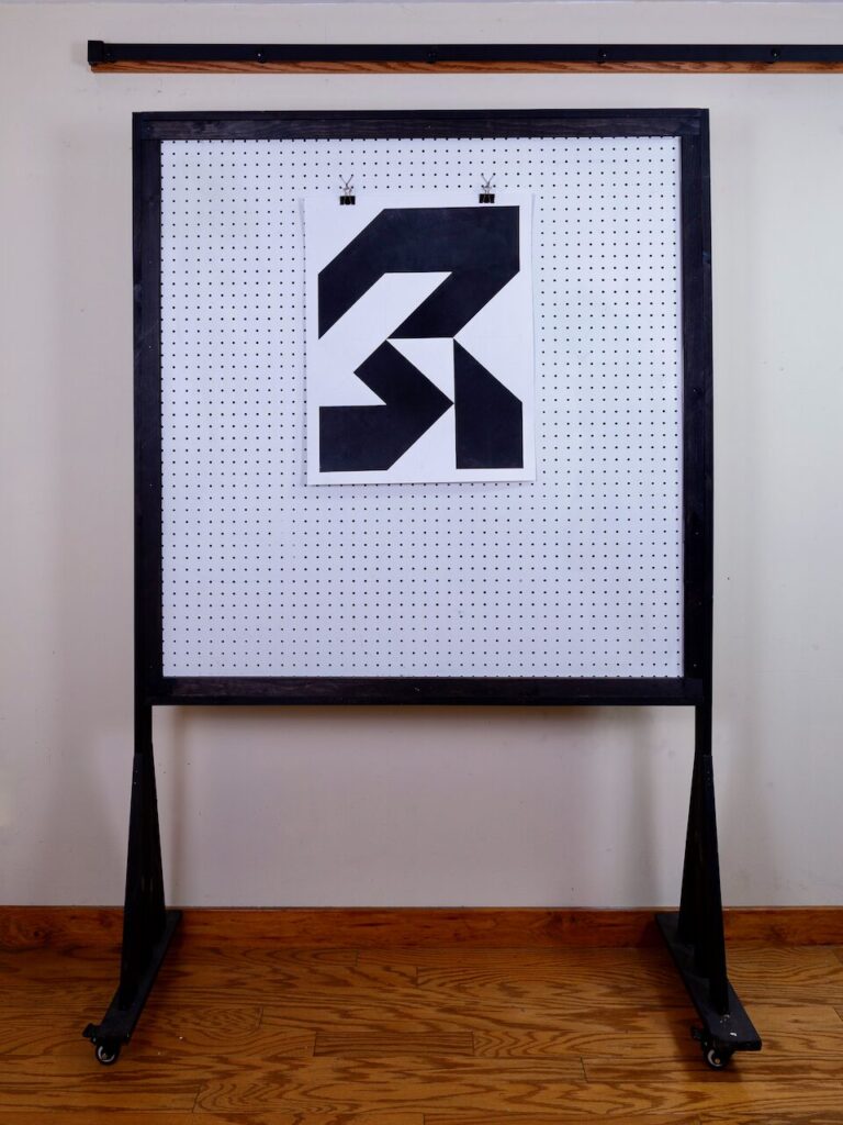 Studio shot of Unnamed (Glyph Drawing No. 14) mounted on pegboard, showing scale of the black-and-white geometric drawing with balanced forms and controlled surface texture.