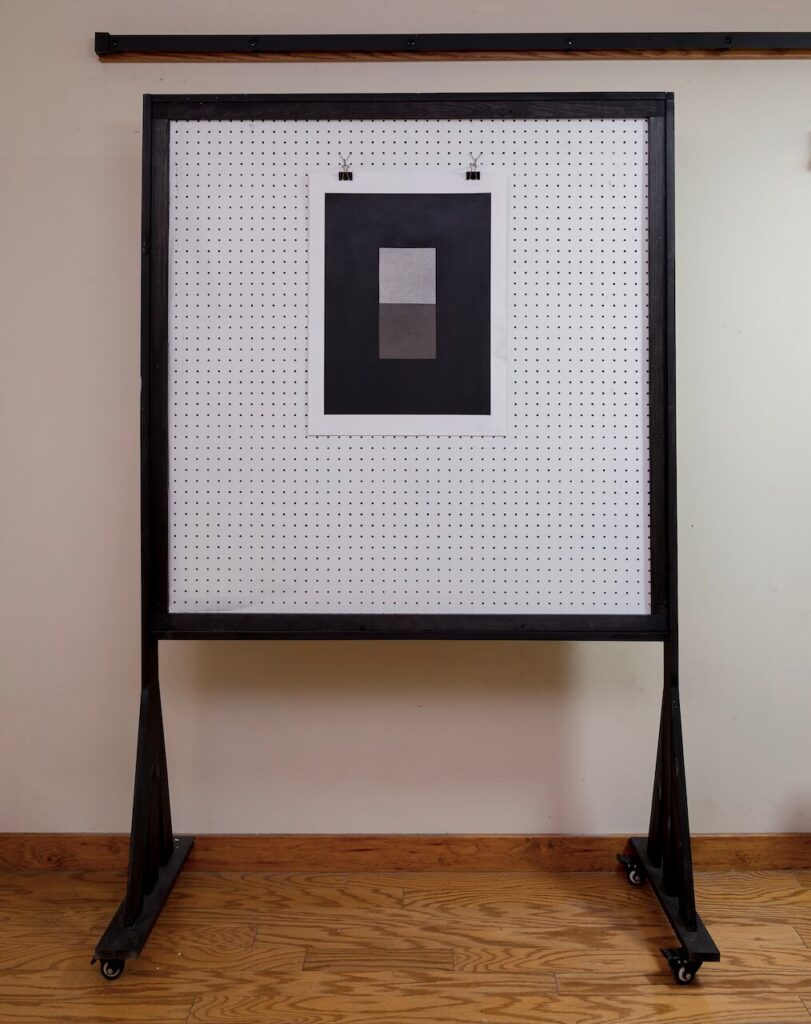 Studio view of Rectangles and Squares, No. 2, showing the 19 x 24" drawing on paper displayed on a pegboard wall, providing scale and context. The composition features a deep charcoal ground, 3×4 grid, and two central squares forming a secondary rectangle.