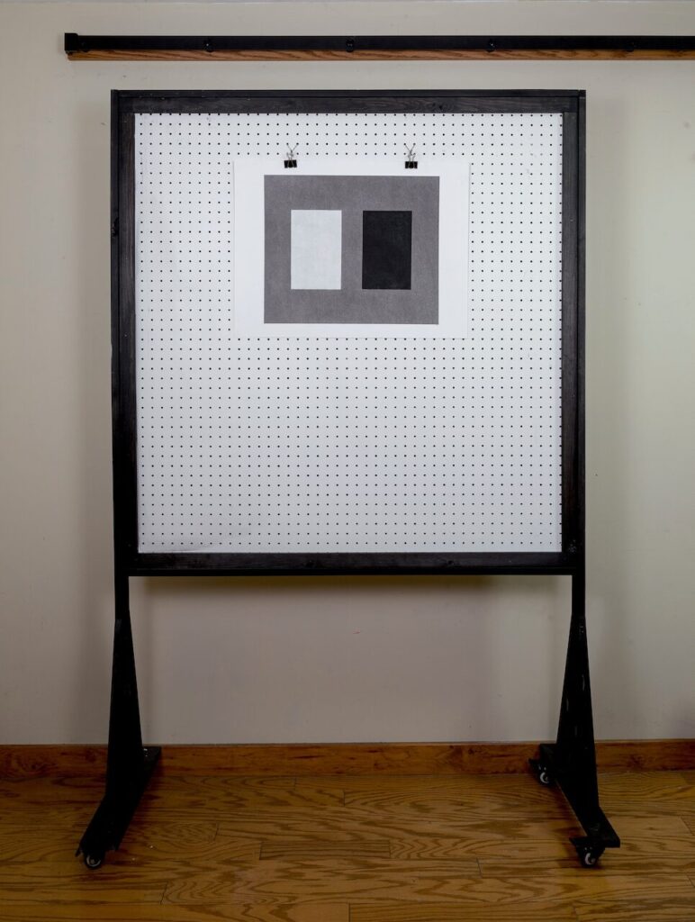 Studio view of Rectangles, No. 1, 18 x 24" drawing displayed on pegboard wall. Composition shows a gray ground with two large vertically oriented golden-ratio rectangles — a light graphite gray on the left and a black matte charcoal on the right.